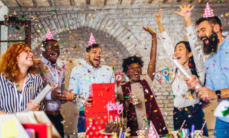 Big group of people celebrating a birthday. There are 6 people in total, all cheering and throwing confetti in the air. Three are wearing party hats. In front of them is a table with birthday decorations, such as a chocolate cake and drink cups.