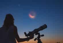 A woman with her back to the camera looks at the crescent moon and two planets through a telescope at sunset