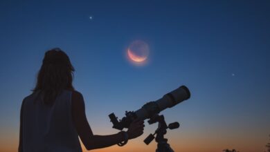 A woman with her back to the camera looks at the crescent moon and two planets through a telescope at sunset