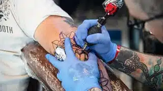 Foto de un tatuador con guantes usando una pistola de tatuar para trazar diseños en el brazo desnudo de una persona.