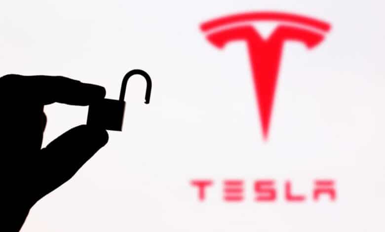 A hand holding an unlocked padlock in front of a blurry tesla logo.